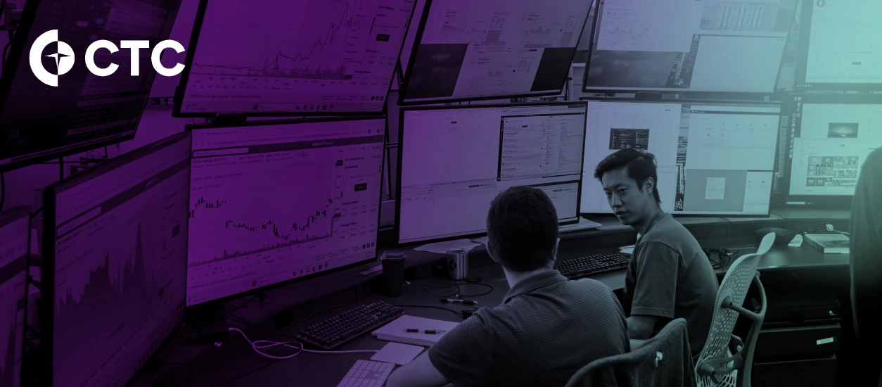 Two coworkers talk at a desk in front of monitors showing stock charts. A CTC logo is in the upper left corner.